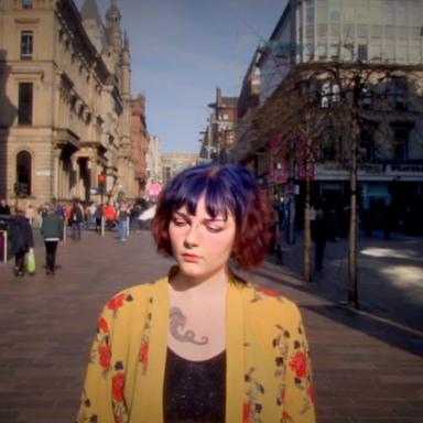 Young person with short blue and red hair, with tattoos and piercings, wearing a yellow patterned kimono and a black top.