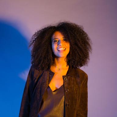 Headshot of young person with curly hair, wearing hoop earrings, a black jacket and grey top.