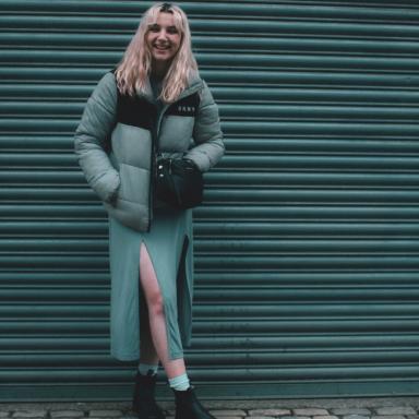 Young person with wavy blonde hair, wearing a grey maxi dress with a grey puffy jacket. She is standing with her legs crossed and smiling at the camera.