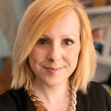 Woman with blonde shoulder length hair wearing black top and gold chain smiles at camera