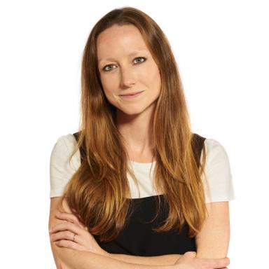 Woman with long strawberry blonde hair wearing black pinafore dress and white t-shirt poses with crossed arms