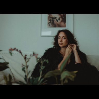 Kasia sitting on a sofa wearing a fuzzy black coat, with houseplant in foreground