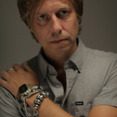 Man with blonde hair wears grey shirt and faces camera with left arm rested across their chest