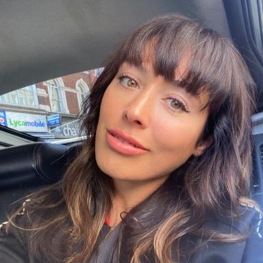 Martha has brown long hair with a fringe and smiles at camera as she sits in car