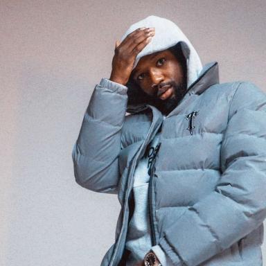 Man wearing light blue puffer jacket and grey hoodie stands facing camera with right hand rested on head