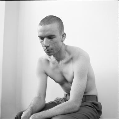 Joe Winter shot topless in black and white, they are sitting on a stool and leaning forward