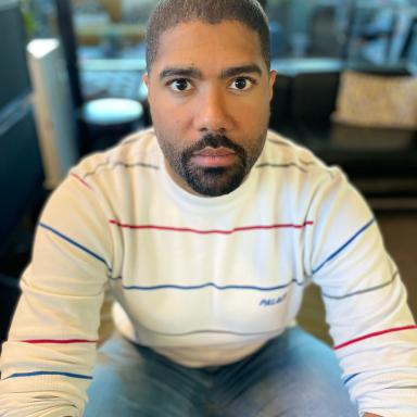 Man with short shaved hair wearing white jumper with coloured stripes and blue jeans faces camera