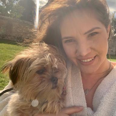 Woman with nose piercing and white cardigan holding a cute tiny brown dog with shaggy fur, outside on a sunny day