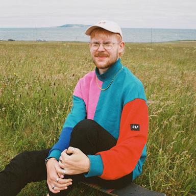 sweet philly sits on a skateboard on the grass and wears a colourful jumper