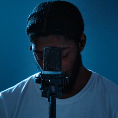 a man wearing a black beanie looks down. a microphone is in front of him. the background is blue