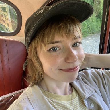 A woman wearing a cap and shirt smiling into the camera. Pale with freckles. 