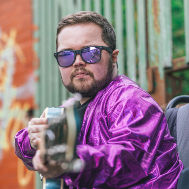 alex james wears a shiny purple jacket, sunglasses and plays a guitar while looking at the camera