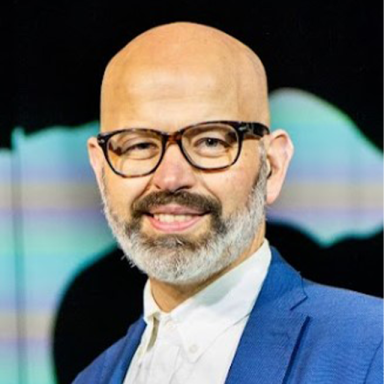 a bald man with black glasses wears a white shirt and blue blazer