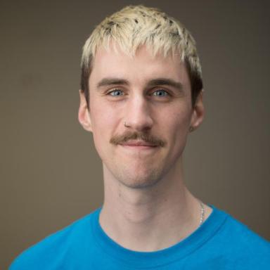 a man with bleached blonde hair and a brown moustache wears a blue t-shirt