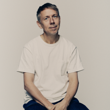 a man with grey short hair sits on a stool. he is wearing a white t-shirt and dark trousers