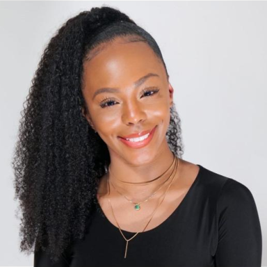 a woman with her curly hair in a ponytail wears a black top, layered necklaces and smiles with her head slightly tilted