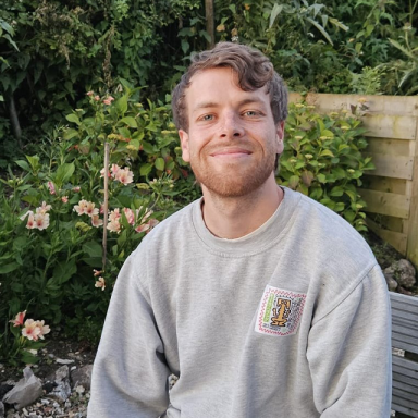 a man with wavy brown hair wears a grey jumper and smiles in a garden with a flower bush