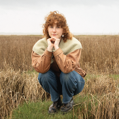 a person with auburn curly hair crouches in a field of dry grass