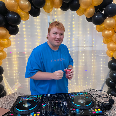 a man in a blue t shirt plays a dj set surrounded by an arch of yellow and black balloons