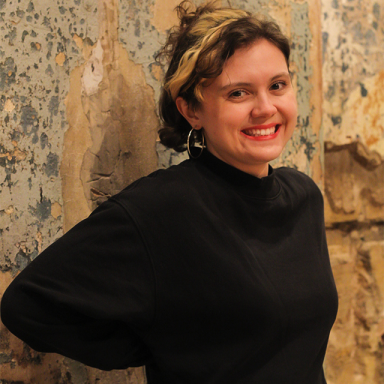 a woman with short brown curly hair smiles and wears a black turtleneck jumper and a headband