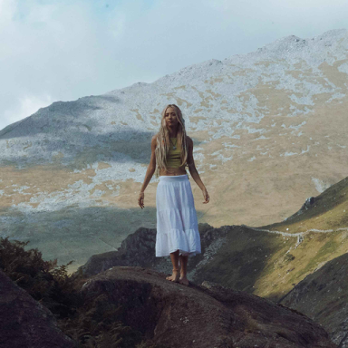 a woman with long blonde hair stands on a hill in the mountains