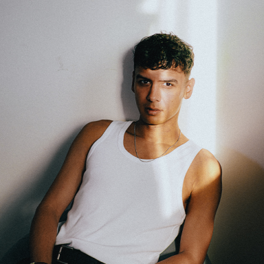 noah bouchard wears a white tank top and reclines; a beam of light hits his face