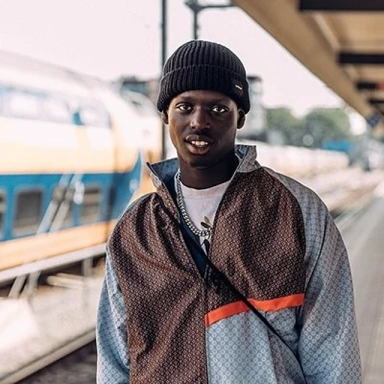 TEEDEMBA is at a train station and wears a black beanie, zip up jacket and cross body bag