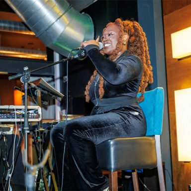 a woman with auburn braids sings into a mic while sitting in a chair on stage